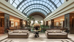 Un hall d’entrée spacieux, doté d’une haute verrière, d’un sol en marbre et de deux canapés placés face à une table centrale agrémentée d’un arrangement floral.