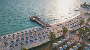 Une vue aérienne d’une plage avec des rangées de transats et de parasols, une jetée qui s’avance dans la mer et un bâtiment à la façade blanche.