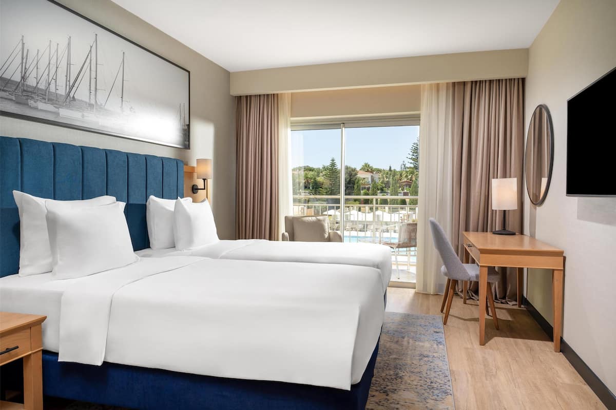Ein Hotelzimmer mit einem großen Bett, einem Schreibtisch, einem Sessel, einem Fernseher und Blick auf einen Pool und Grünflächen.