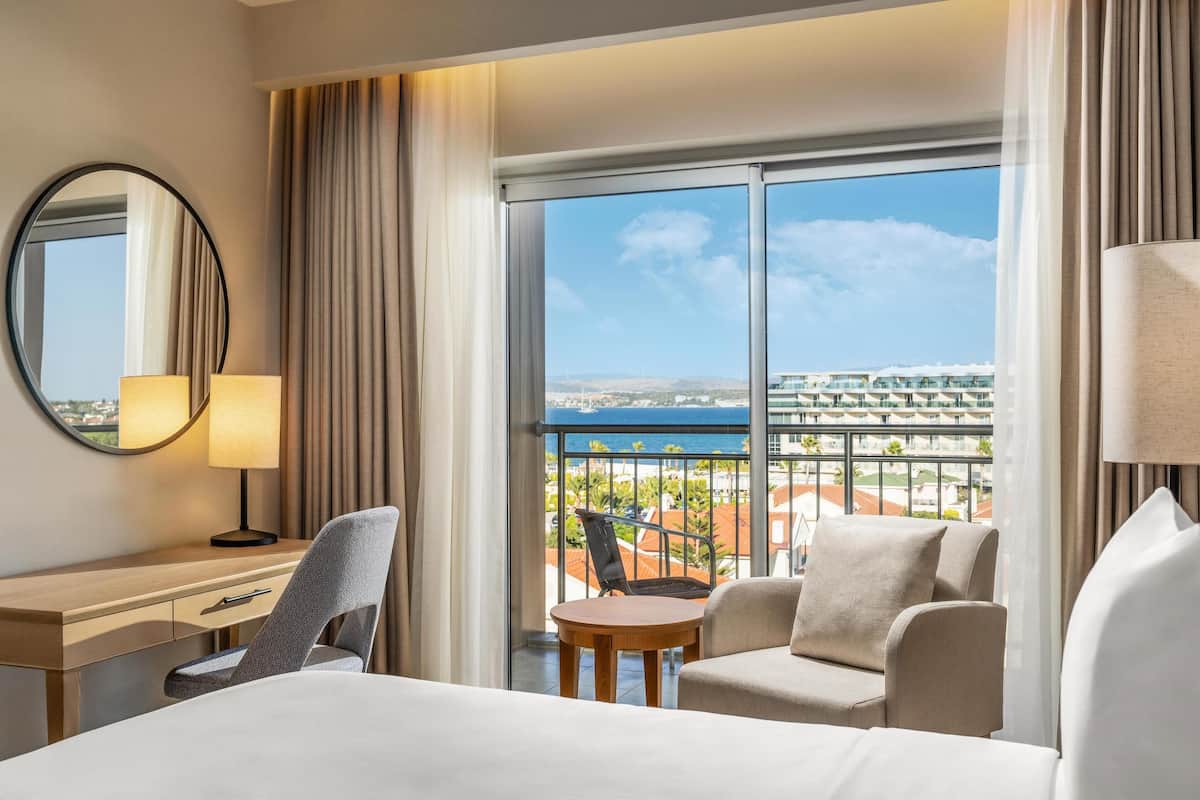 Ein Hotelzimmer mit Bett, Schreibtisch, Stuhl, rundem Tisch und einem Balkon mit Blick auf die Stadt und das Wasser.
