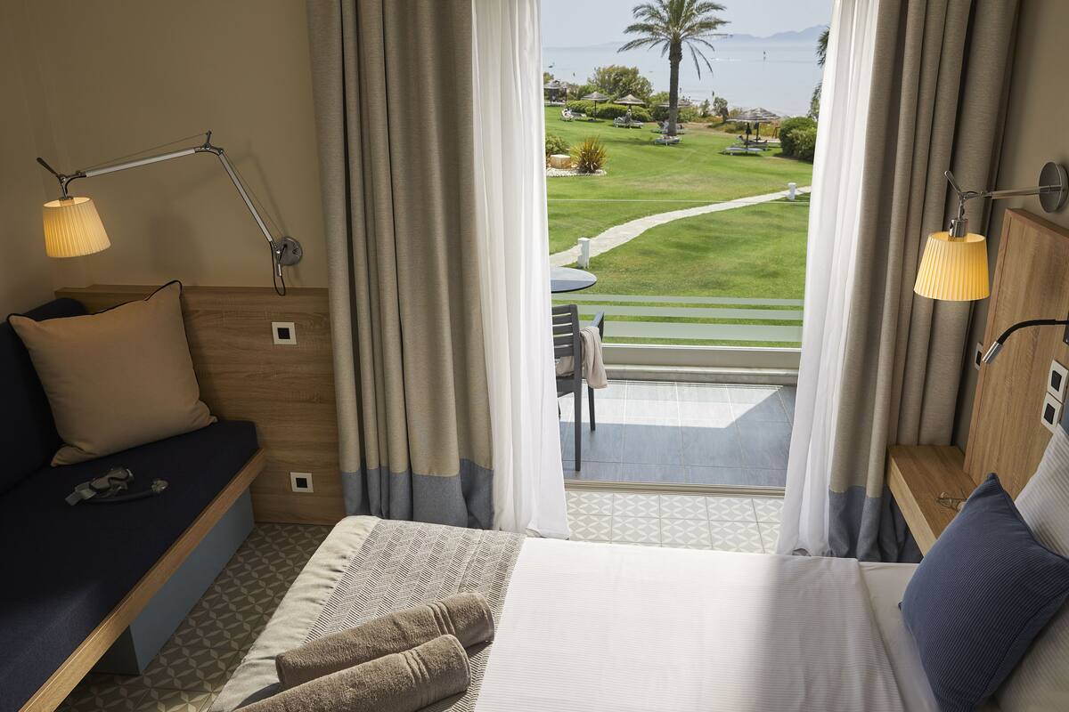 Habitación de hotel con cama, sofá, escritorio, lámpara y vista a un jardín y palmeras.