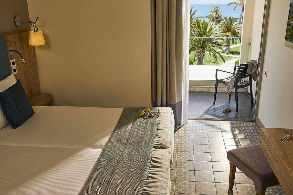 Habitación de hotel con cama, escritorio y vista a palmeras y al océano.