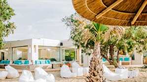 A beachside lounge area with white bean bags, a palm tree, and a modern building with large windows and outdoor seating. - ROBINSON KYLLINI BEACH (Andravida-Kyllini)