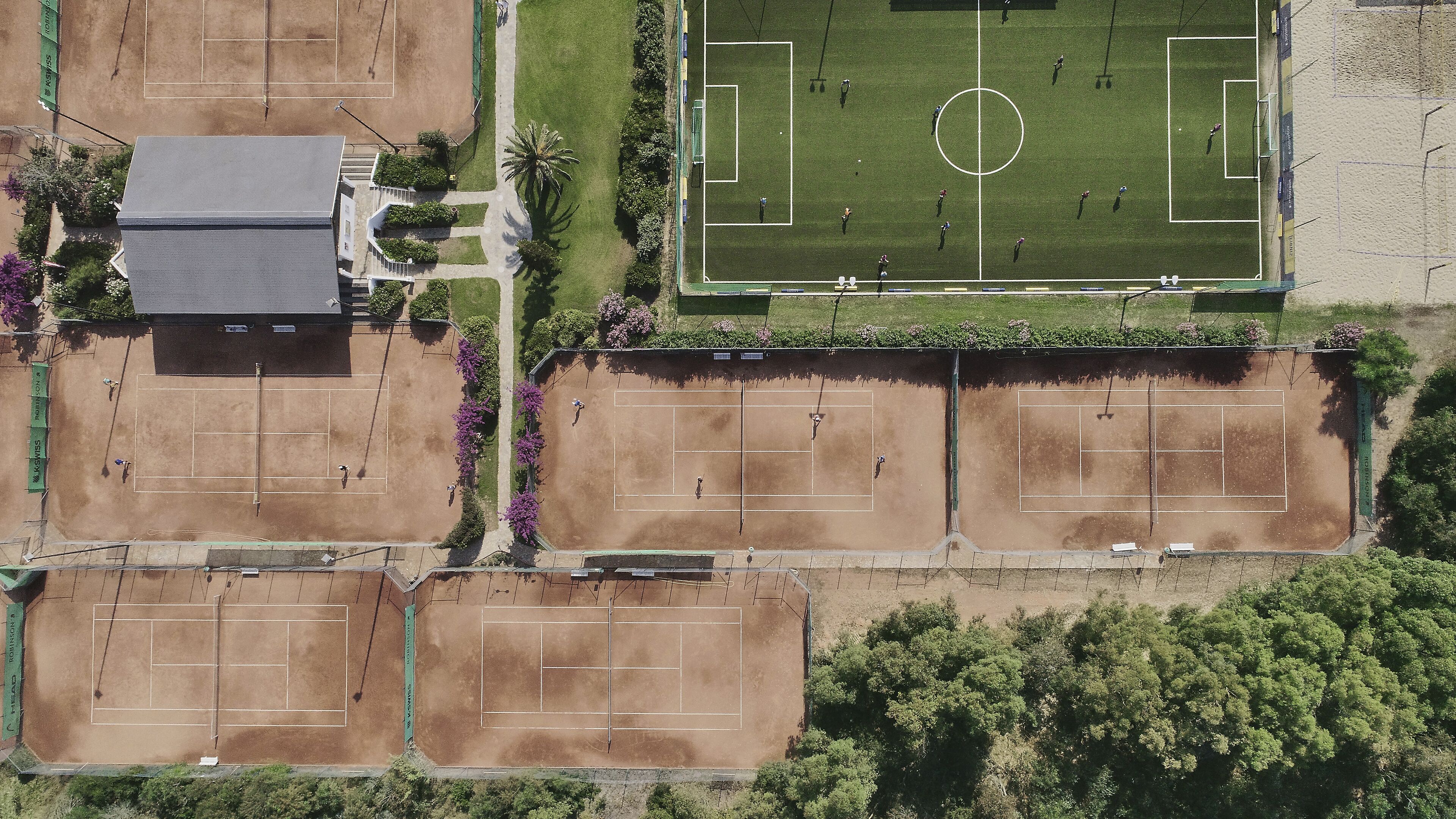 Vue aérienne d’un complexe sportif comprenant plusieurs courts de tennis, un terrain de football et un bâtiment doté d’un toit en pente.