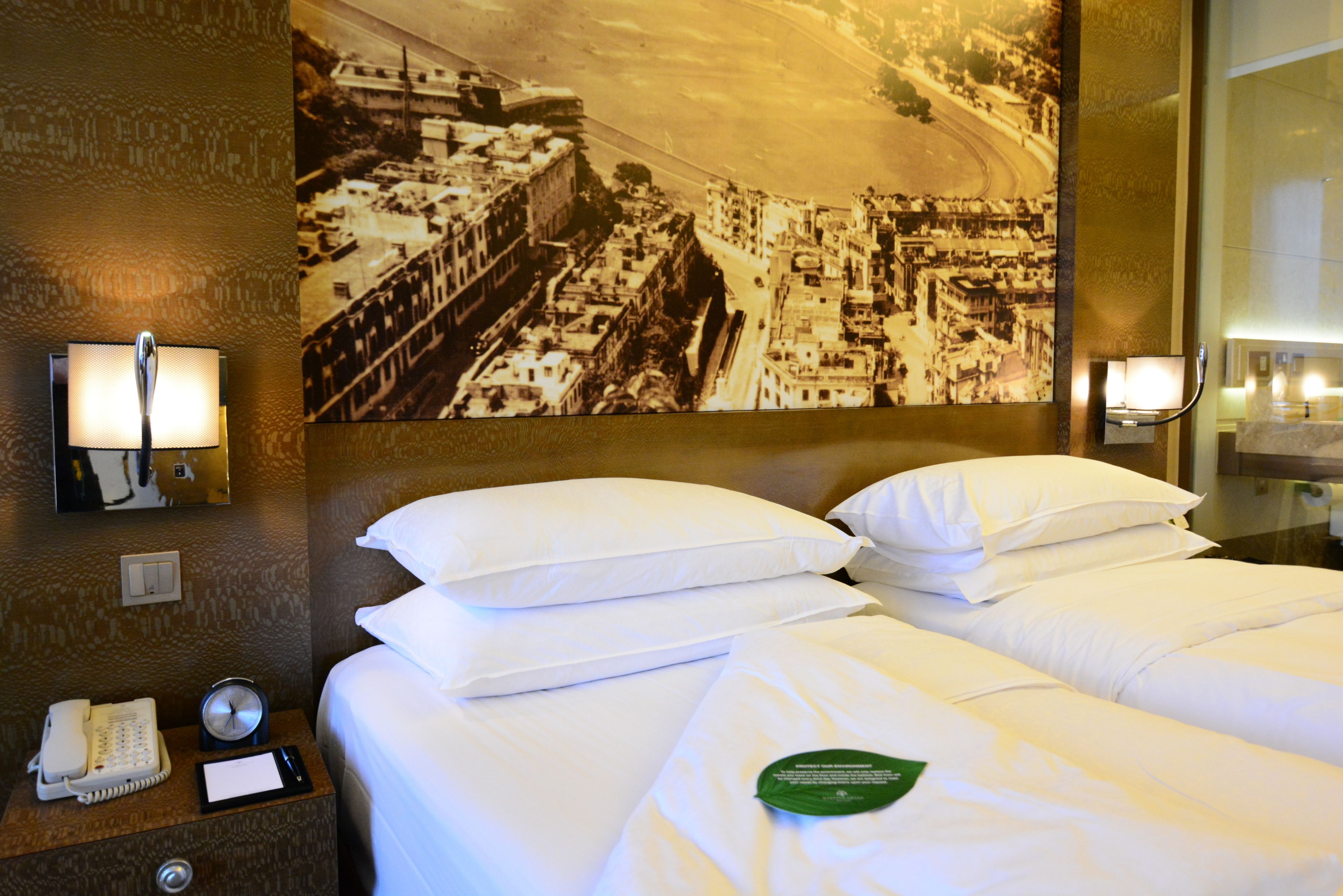 A hotel room with a large bed, two pillows, a bedside table with a phone and a notepad, a wall clock, and a framed historical photograph.