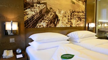 A hotel room with a large bed, two pillows, a bedside table with a phone and a notepad, a wall clock, and a framed historical photograph.