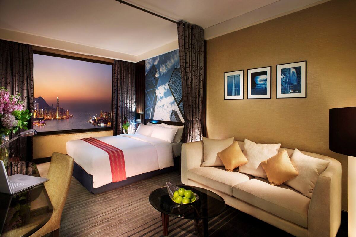 A hotel room with a bed, a sofa, a desk with a laptop, a coffee table with apples, and a large window with a cityscape view.