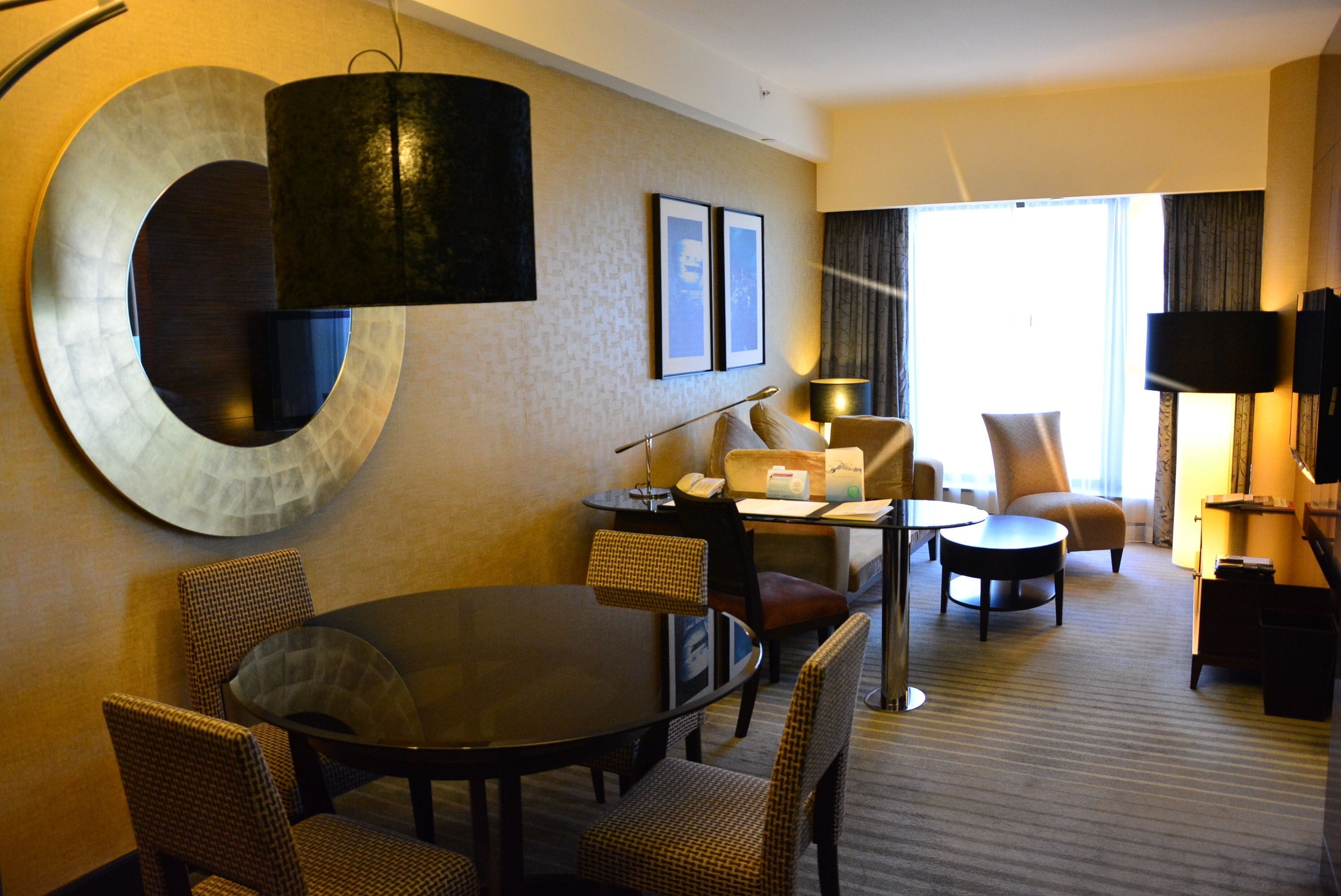 A hotel room with a round dining table, two armchairs, a sofa, a TV, and a large window with curtains.