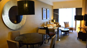 A hotel room with a round dining table, two armchairs, a sofa, a TV, and a large window with curtains.