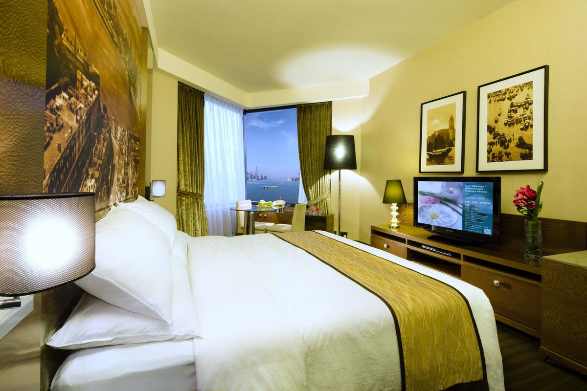 A hotel room with a large bed, a desk with a computer, a TV, and a view of the cityscape through the window.