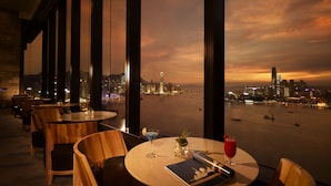 A restaurant with a view of a city skyline at sunset. Tables are arranged with chairs, and there are drinks on the tables. - Harbour Grand Hong Kong (Hong Kong)