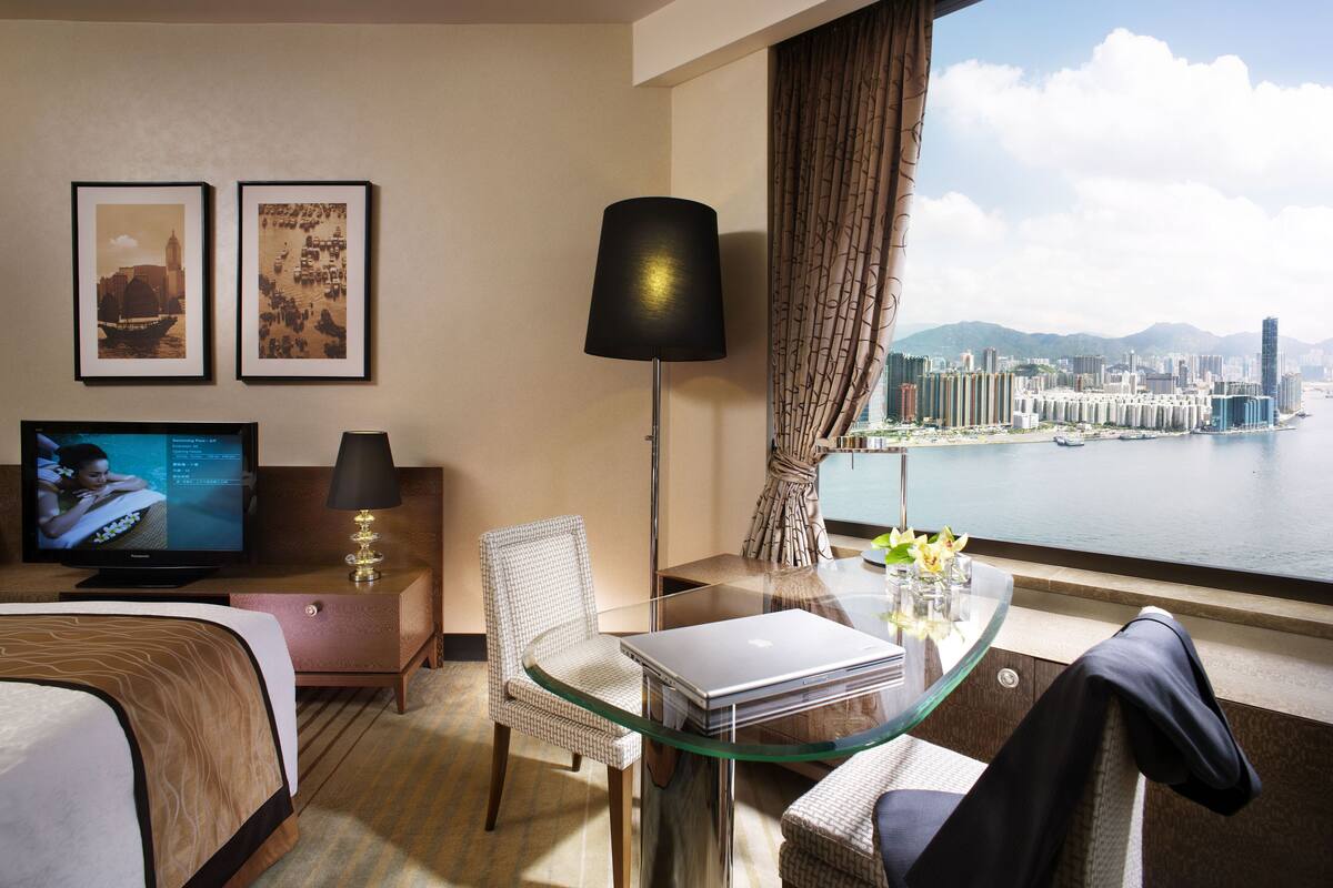 A hotel room with a bed, a desk with a laptop, a chair, a TV, and a view of a cityscape and water.