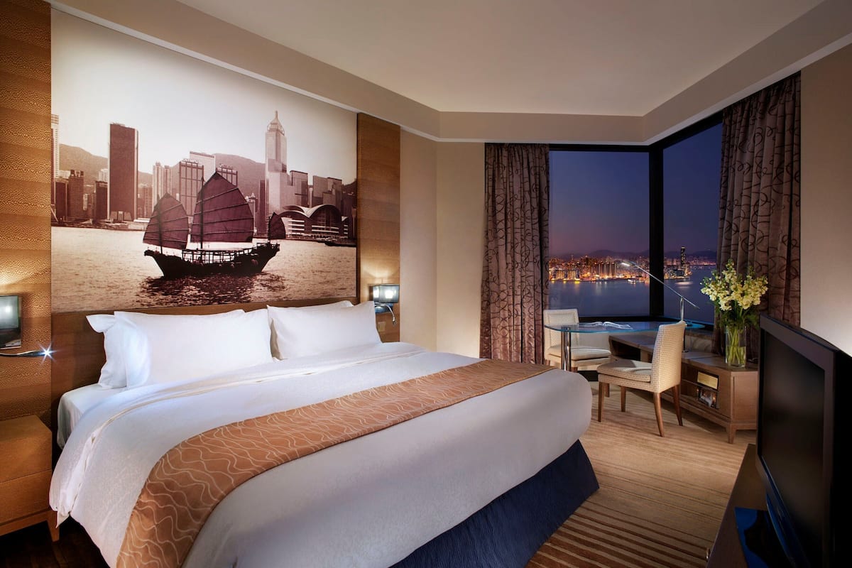A hotel room with a large bed, a desk with a chair, a TV, and a view of a cityscape at night.