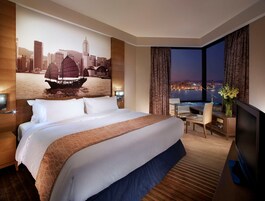 Habitación de hotel con una cama grande, un escritorio con silla, televisión y vista a un paisaje urbano nocturno.