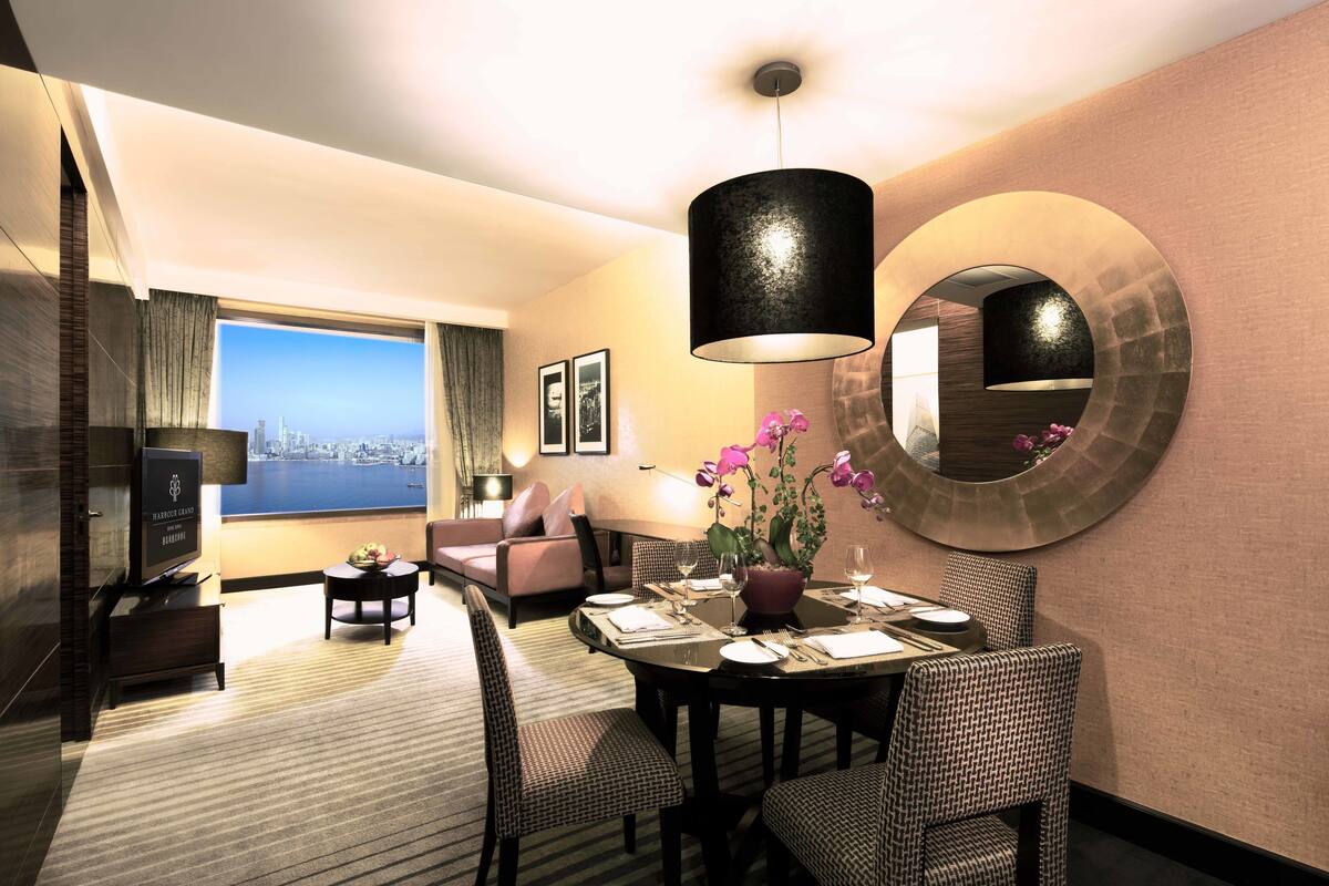 A hotel room with a dining area, a large window with a city view, a television, and a potted plant with flowers.