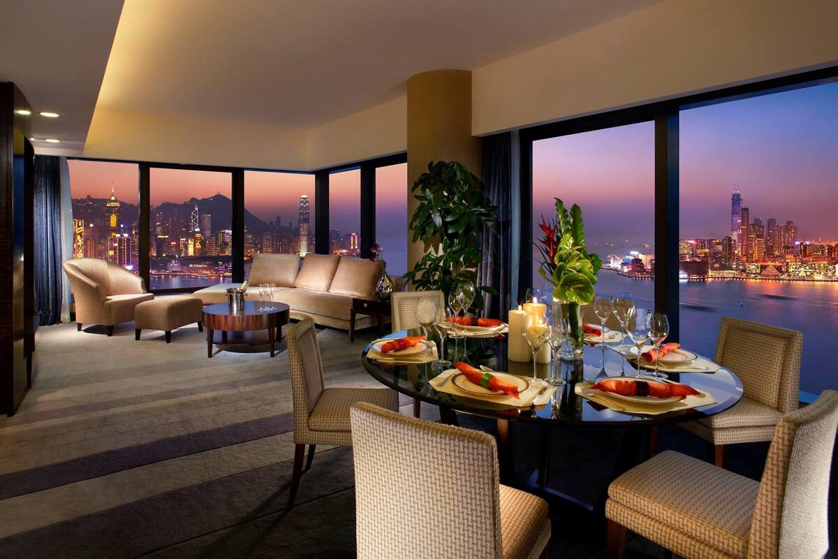 A modern hotel room with a city view, a dining area with a table set for two, and a cozy seating area with a sofa and armchairs.