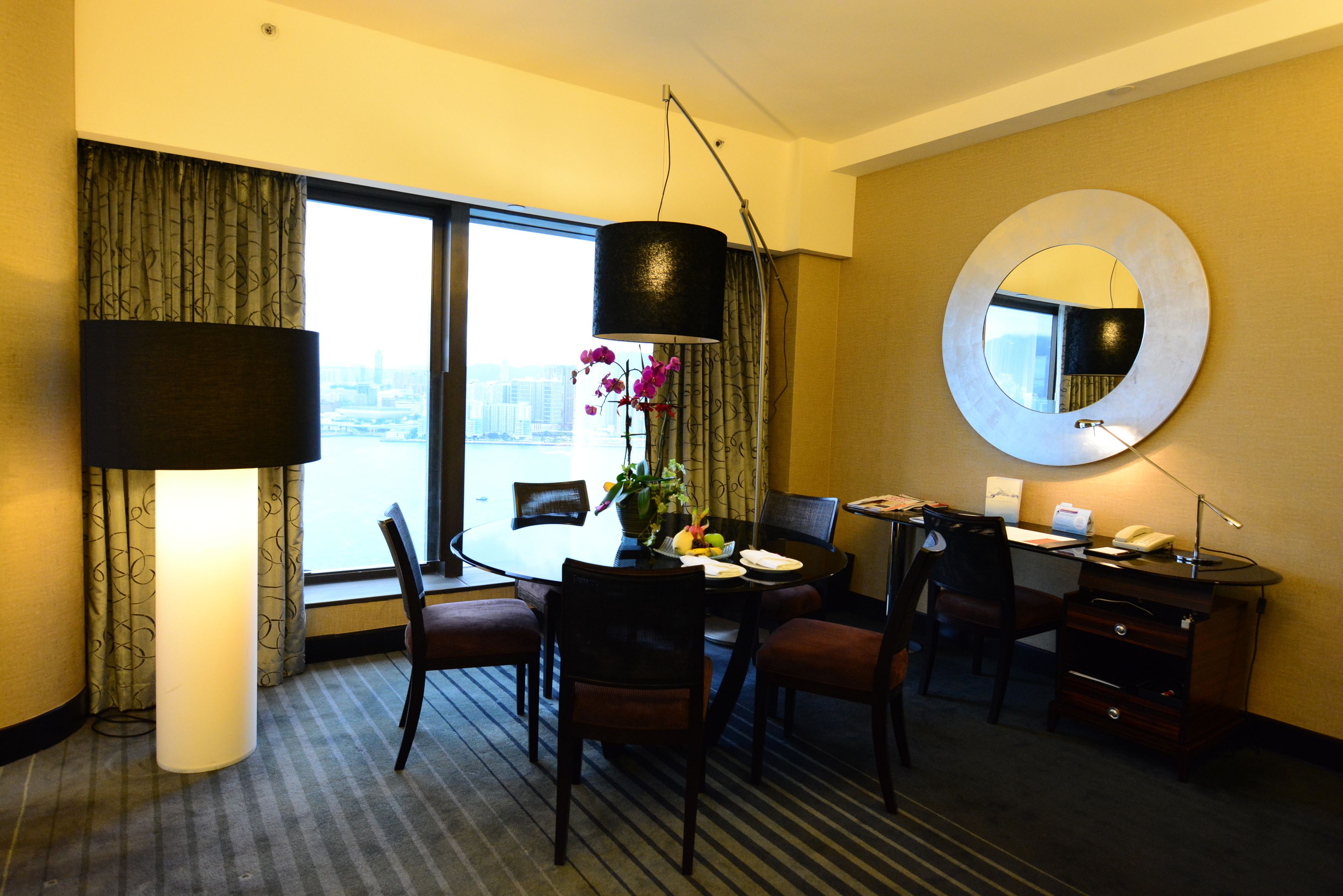 A hotel room with a dining table, chairs, a desk with a computer, a lamp, a mirror, and a view of the city through the window.