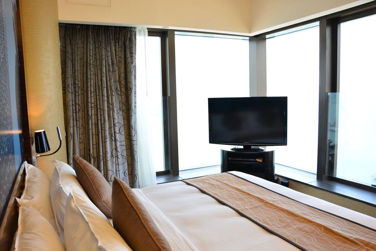 A hotel room with a large bed, a flat-screen TV on a stand, a window with curtains, and a view of the city.