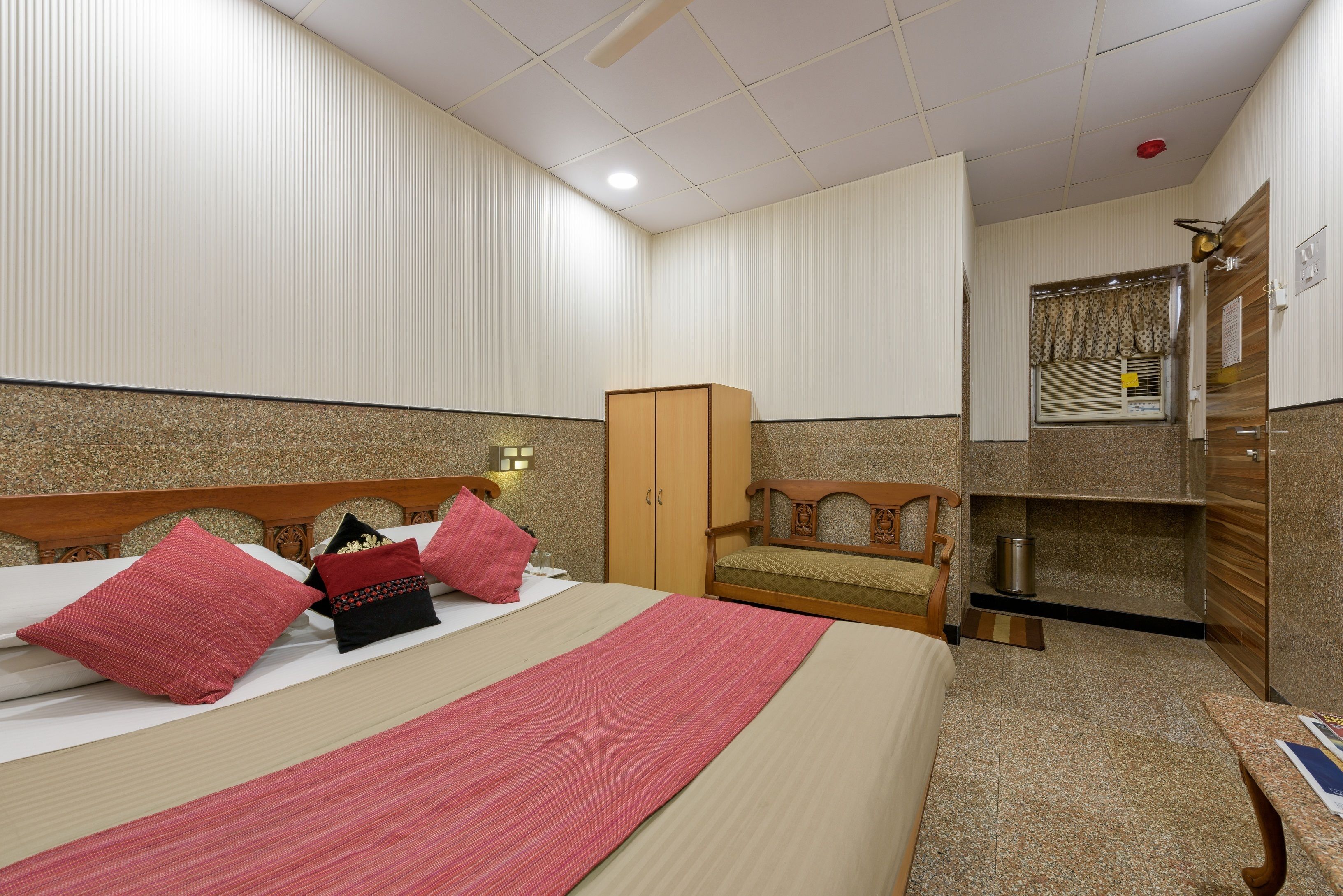 A hotel room with a bed, a wooden wardrobe, a small wooden chair, a desk, and a window with curtains.