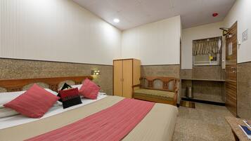 A hotel room with a bed, a wooden wardrobe, a small wooden chair, a desk, and a window with curtains.