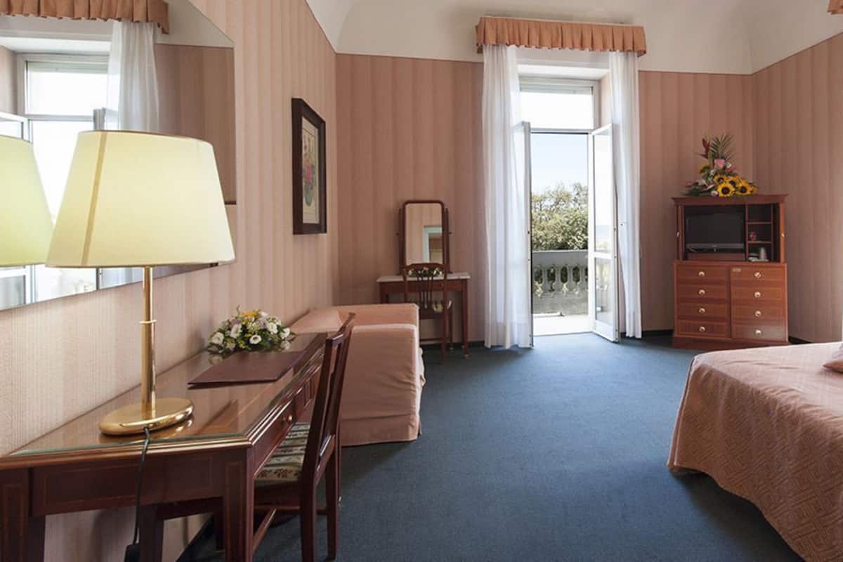 A hotel room with a desk, chair, bed, dresser, and a balcony with a view.