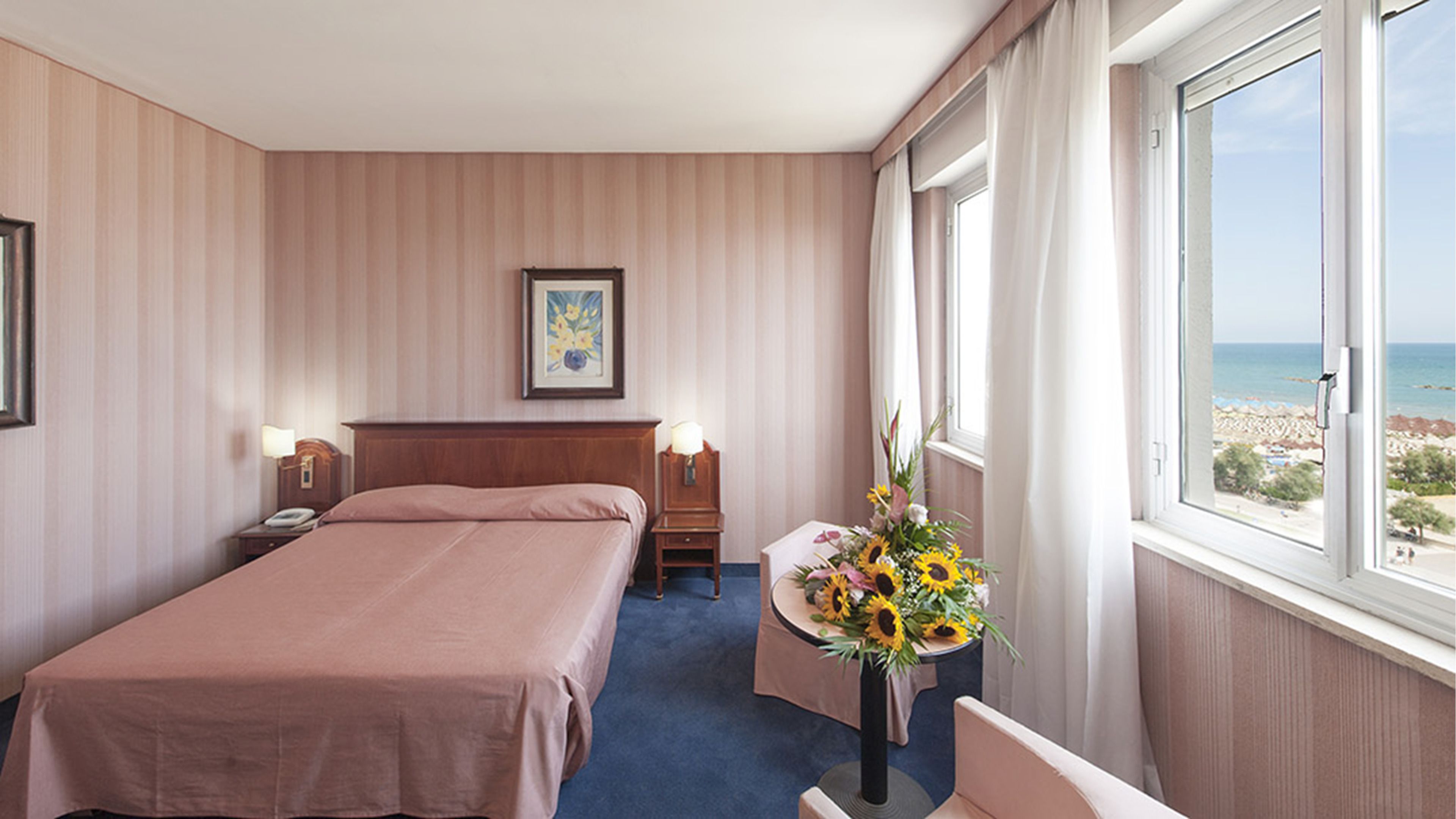 A hotel room with a bed, bedside tables, a desk with a chair, a window with a beach view, and a framed picture on the wall.