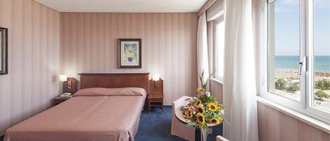 A hotel room with a bed, bedside tables, a desk with a chair, a window with a beach view, and a framed picture on the wall.
