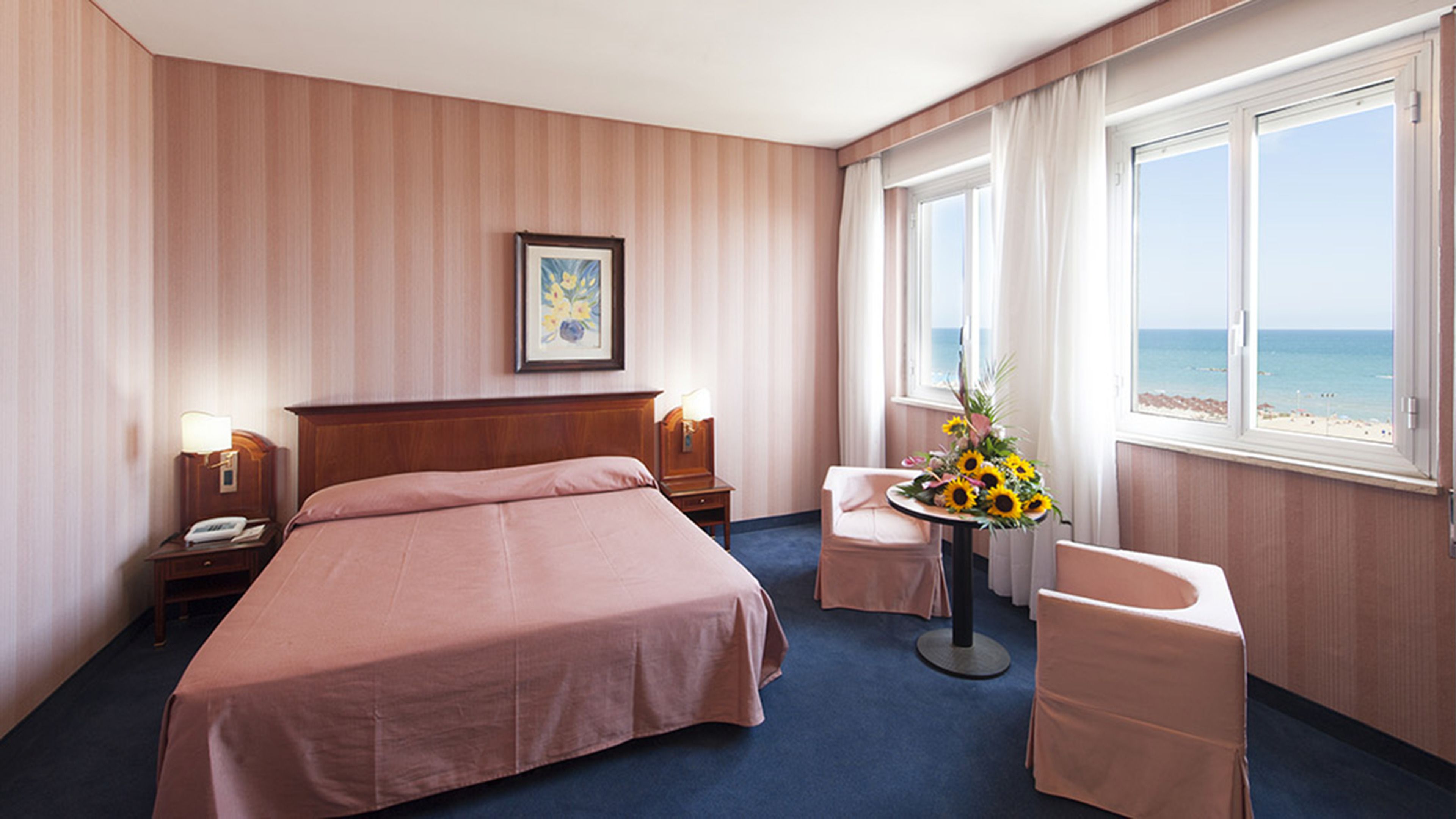 Une chambre d’hôtel avec un grand lit, des tables de chevet avec des lampes, une petite table ornée d’un vase de fleurs, un fauteuil et une vue sur la plage grâce à de grandes fenêtres.