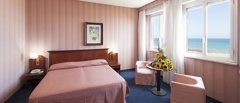 Une chambre d’hôtel avec un grand lit, des tables de chevet avec des lampes, une petite table ornée d’un vase de fleurs, un fauteuil et une vue sur la plage grâce à de grandes fenêtres.