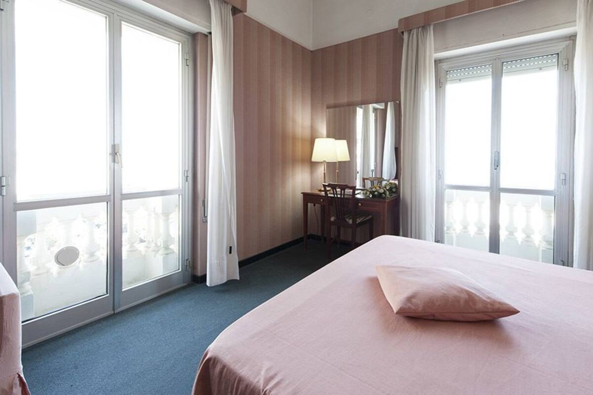 A hotel room with a large bed, a desk with a lamp, and a view of a balcony with white railings.