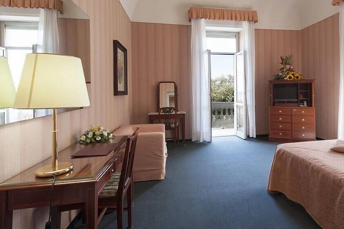 A hotel room with a desk, chair, bed, dresser, and a balcony with a view.