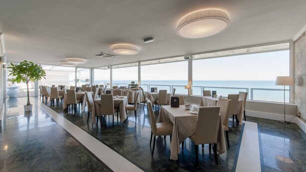 A spacious dining area with tables and chairs, large windows offering a view of the sea, and a potted plant. - Hotel Esplanade (Pescara)