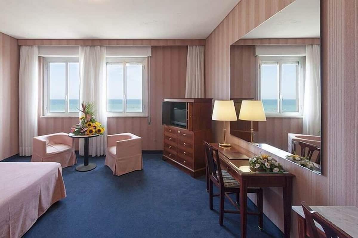 A hotel room with a bed, two armchairs, a small round table with a vase of flowers, a TV, a dresser, and a desk with two lamps.
