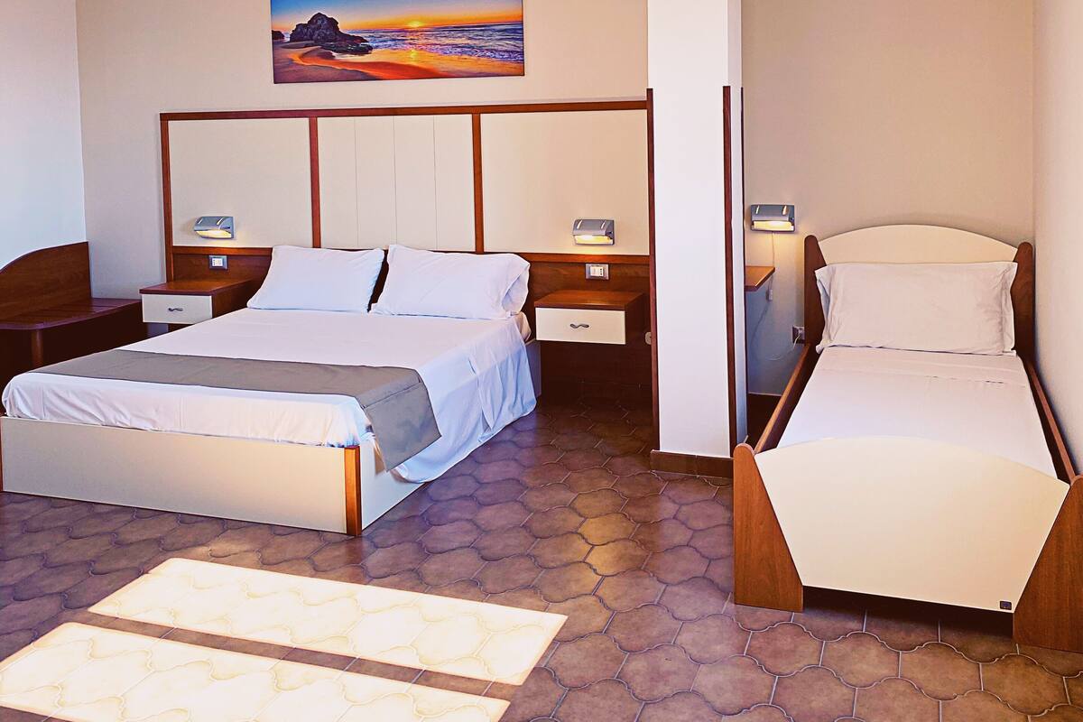 A hotel room with a large bed, a smaller bed, a nightstand, a bedside table, and a painting of a sunset on the wall.