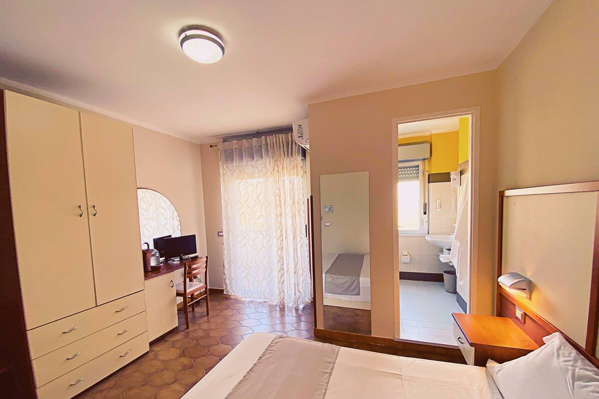 A bedroom with a bed, a desk with a computer, a wardrobe, and a bathroom visible through an open door.