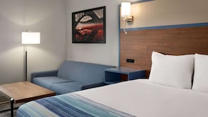 A hotel room with a bed, a desk, a chair, a nightstand, a wall-mounted lamp, and a painting.