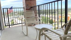 A balcony with two white chairs and a small table, a brick pillar, and a view of a cityscape with a flag.