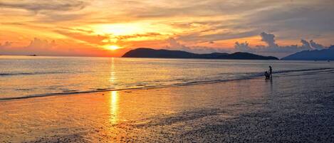 A beach at sunset with calm waters reflecting the sky, a person walking, and distant hills.