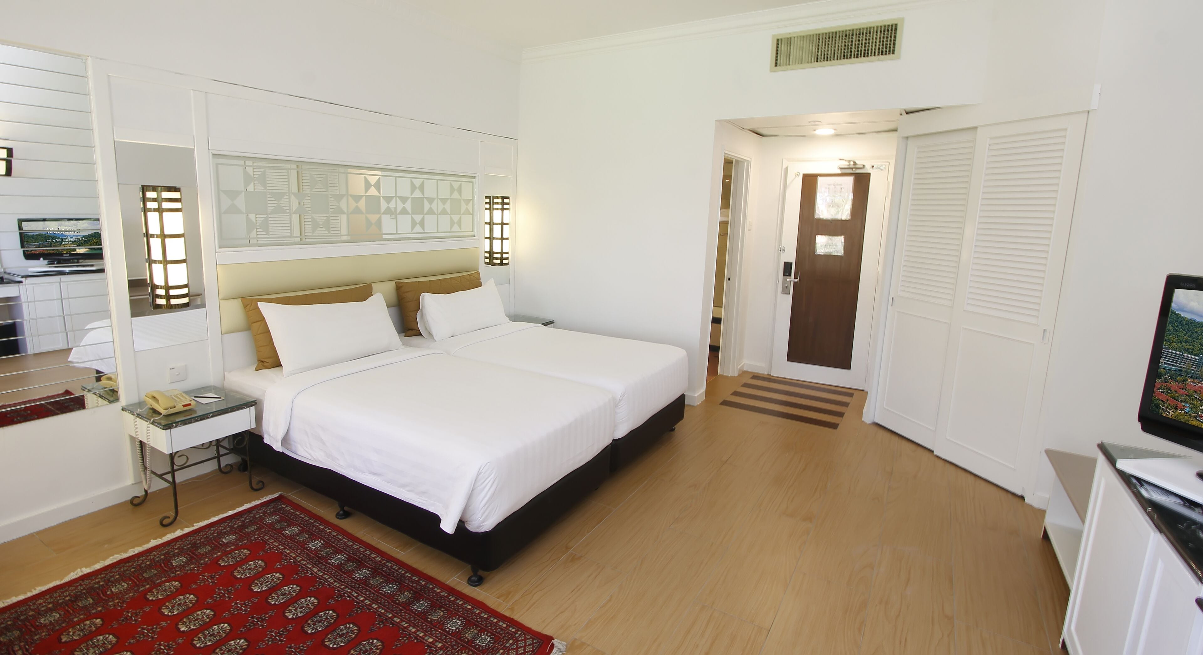 A hotel room with a large bed, a bedside table with a lamp, a red patterned rug, and a television on a cabinet.