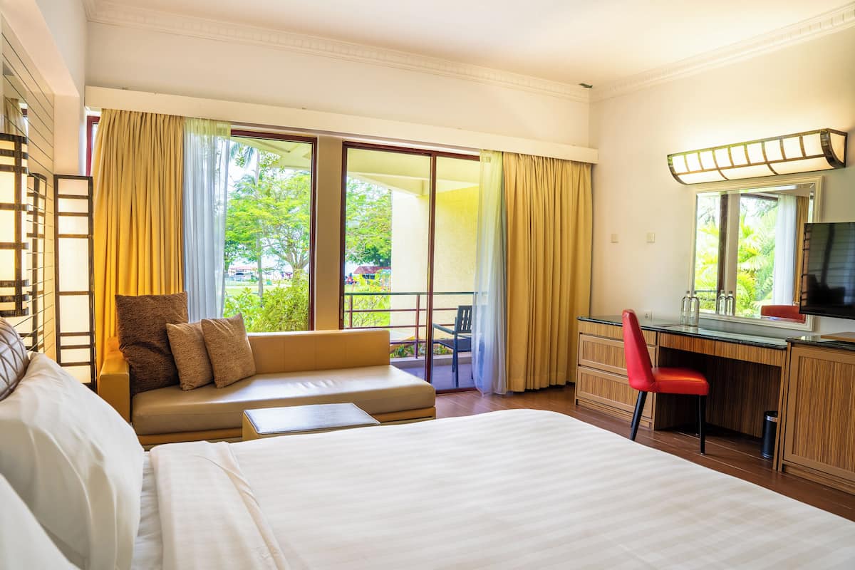 Une chambre d’hôtel avec un lit, un canapé, un bureau et une chaise. Il y a une grande fenêtre avec des rideaux, une vue sur la verdure à l’extérieur et un miroir au-dessus du bureau.