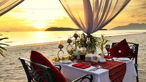 Une table dressée pour deux, installée sur la plage, avec une nappe blanche, des serviettes rouges et des bougies.
