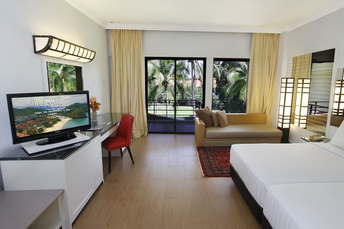 Une chambre d’hôtel avec un lit, un canapé, un téléviseur posé sur un bureau et une vue sur un paysage tropical.