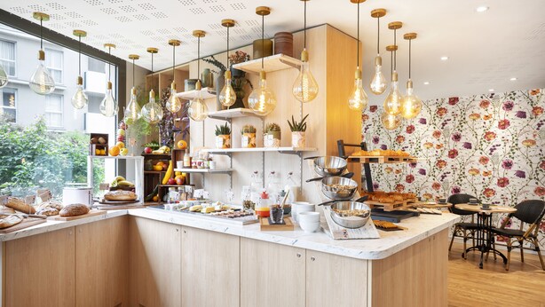 Une cuisine moderne avec des meubles en bois, un îlot central où sont disposés des aliments, des luminaires suspendus et un papier peint à motifs floraux.