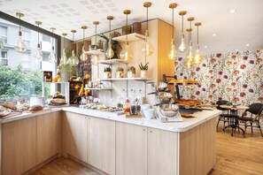 A modern kitchen with wooden cabinets, a central island with food items, hanging light fixtures, and a floral wallpaper. - Séjours & Affaires Paris Malakoff (Malakoff)