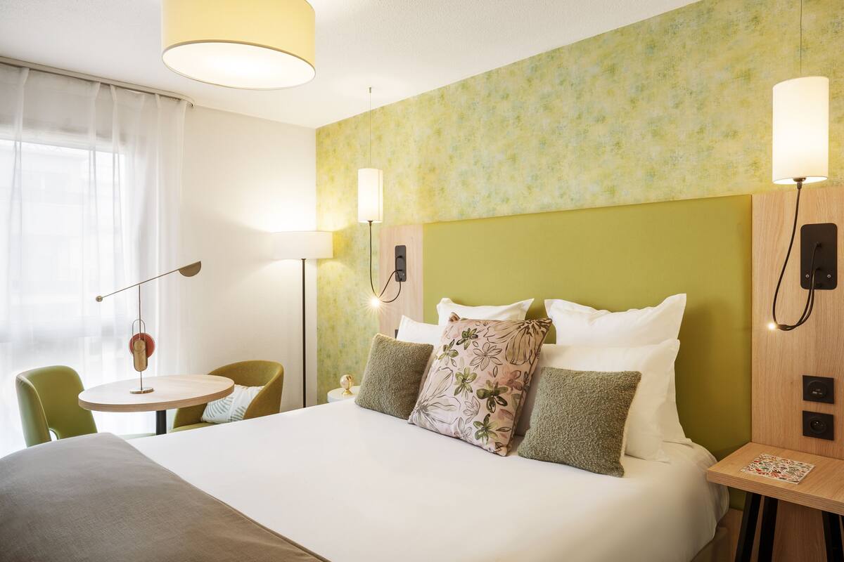 A hotel room with a bed, bedside table, lamp, chair, and a small round table.