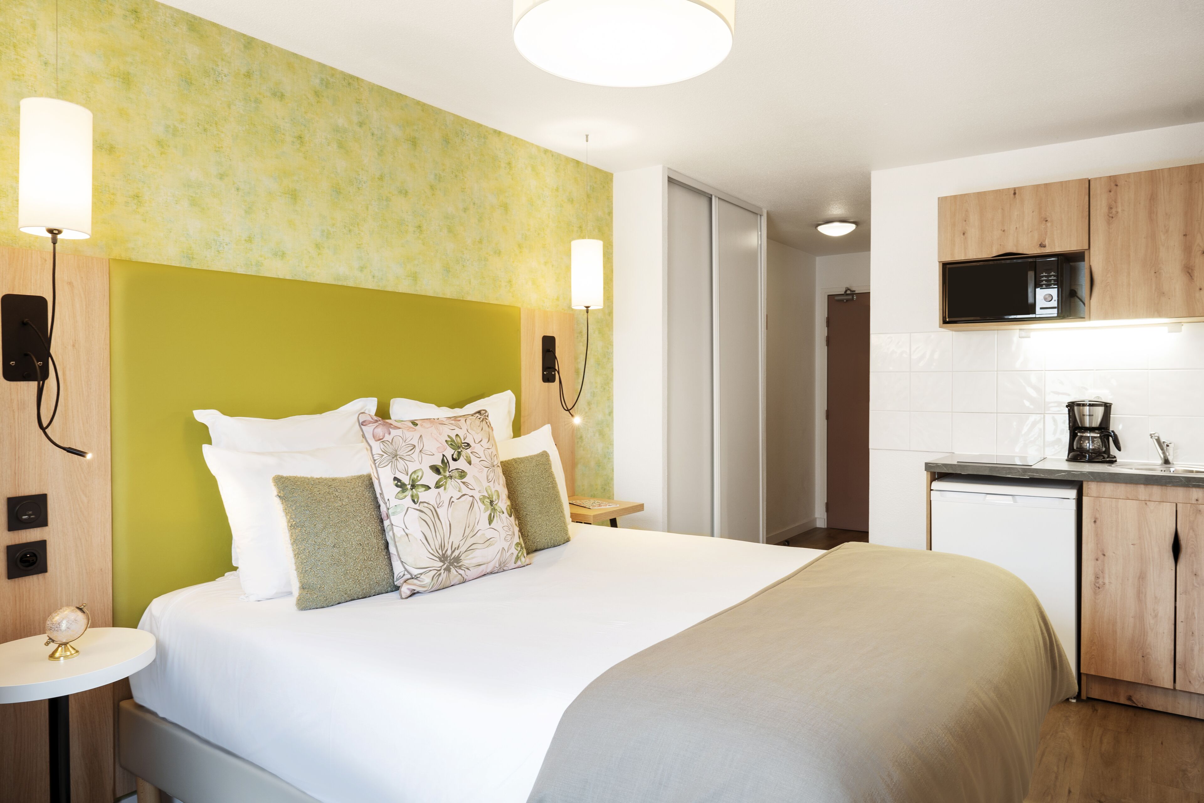 A hotel room with a bed, bedside table, lamp, and a kitchenette area with a microwave.