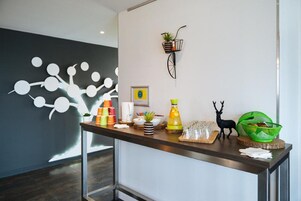 Uma área de recepção com design moderno, incluindo uma obra de arte na parede, um vaso de bicicleta e diversas comodidades como um espaço para café e uma cesta de frutas.