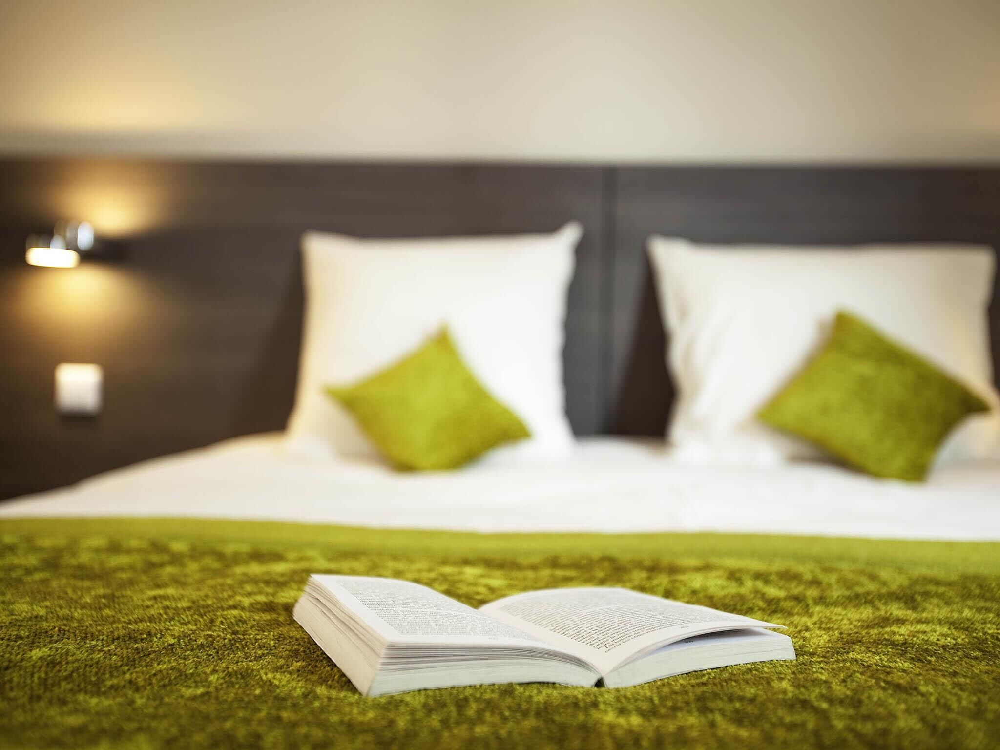 Una cama con ropa de cama blanca y almohadas verdes, un libro abierto sobre una alfombra verde, y un cabecero oscuro.