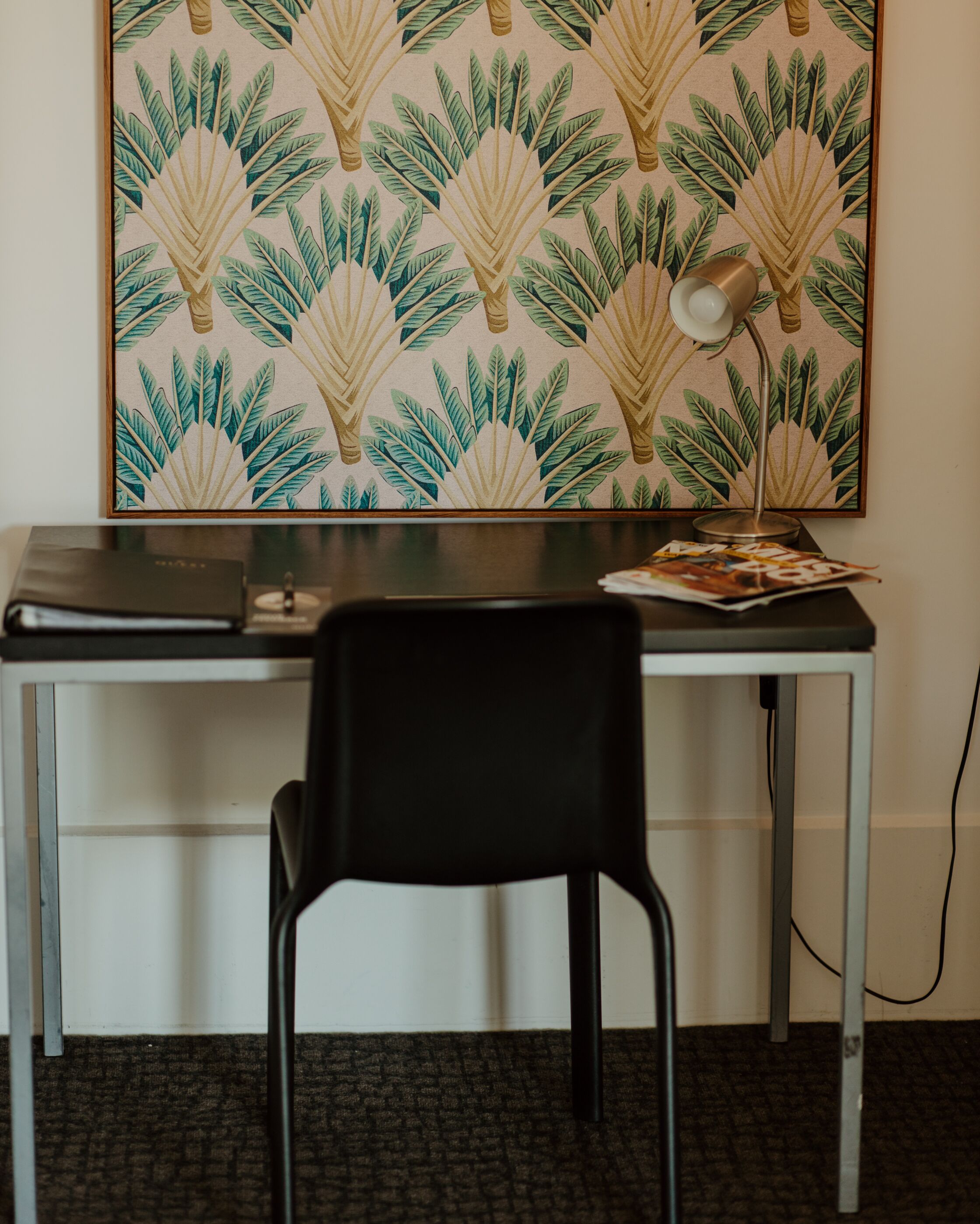 A desk with a laptop, a lamp, and a magazine, set against a wall with a tropical leaf pattern.