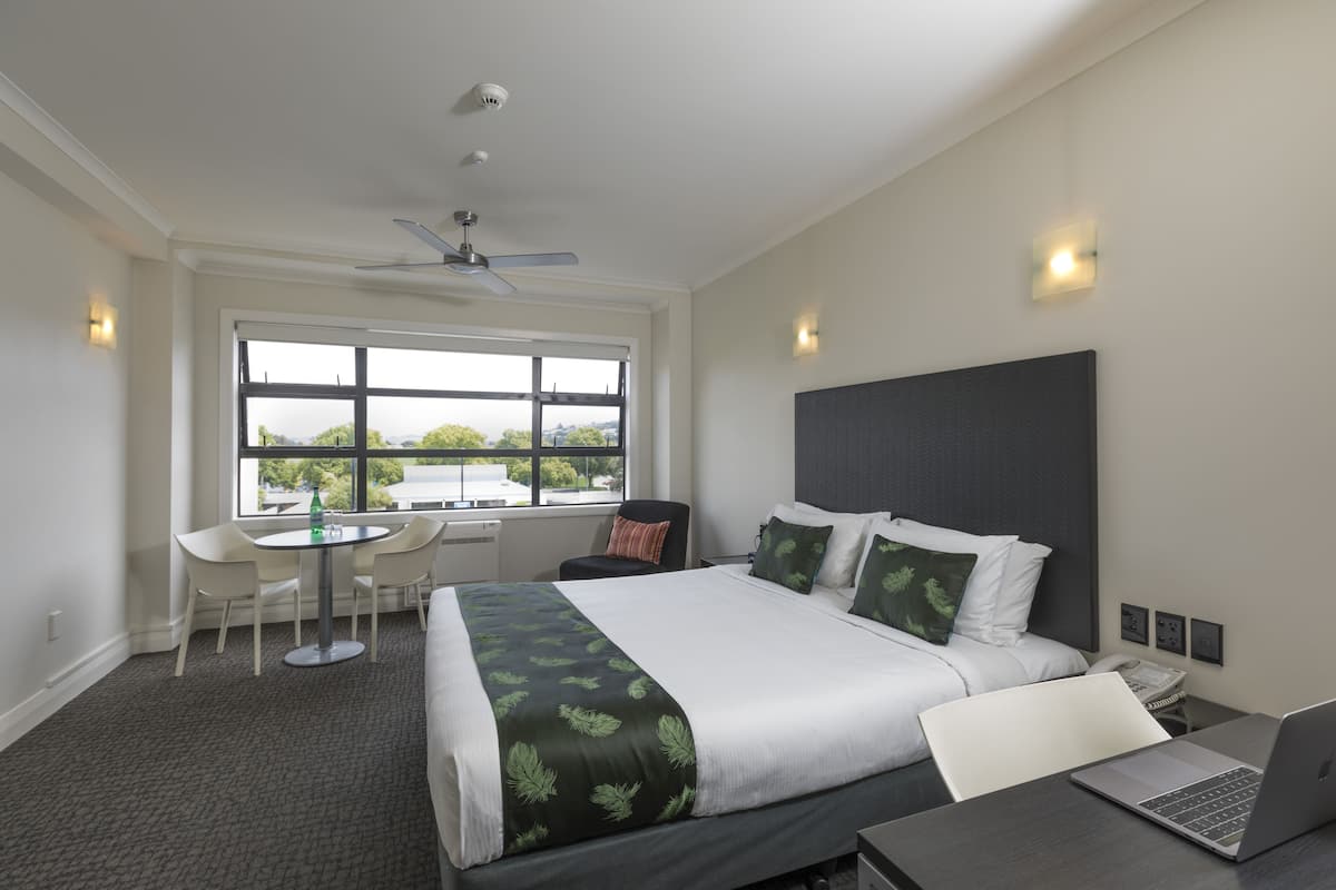 Quarto de hotel moderno com uma cama grande, uma escrivaninha com um laptop, uma mesa de jantar com cadeiras e uma janela com vista para o exterior.